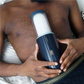 A person lies down holding the SVAKOM Alex Neo Thrusting Masturbating Sleeve, a cylindrical device with a transparent top section and buttons on its body, against a white pillow. App control ensures seamless interactivity. The individual is wearing dark shorts with a light stripe along the waistband.